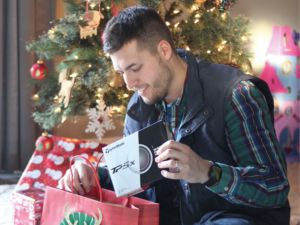 A man sitting on the floor next to a Christmas tree, opening a gift.