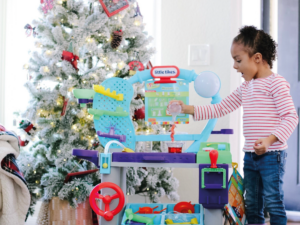 A little girl happily playing with a toy set, with a Christmas tree in the background.