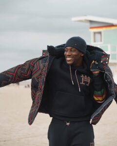 A man wearing a Tommy hoodie and jacket stands on the beach, smiling.