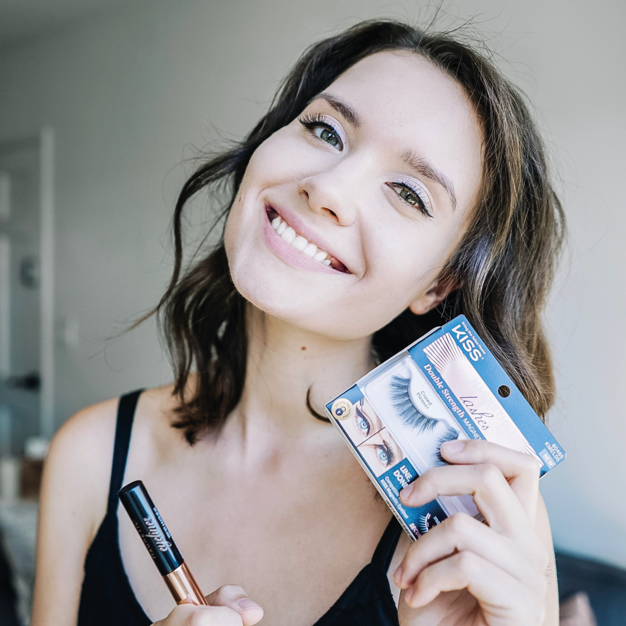 A smiling woman showcasing makeup products from Kiss Cosmetics.