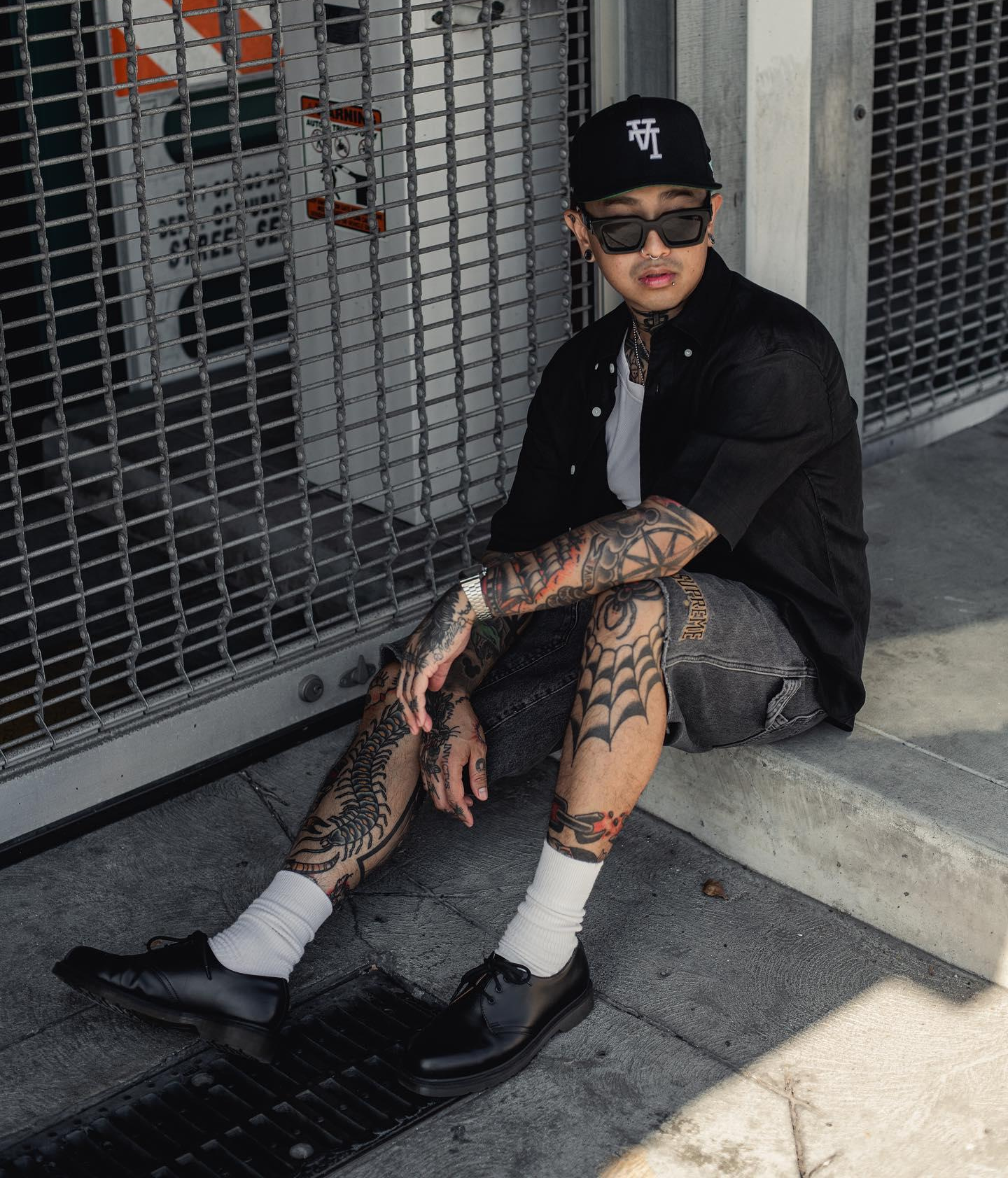 Man with tattoos sitting on the floor, wearing sunglasses and Dr. Martens.