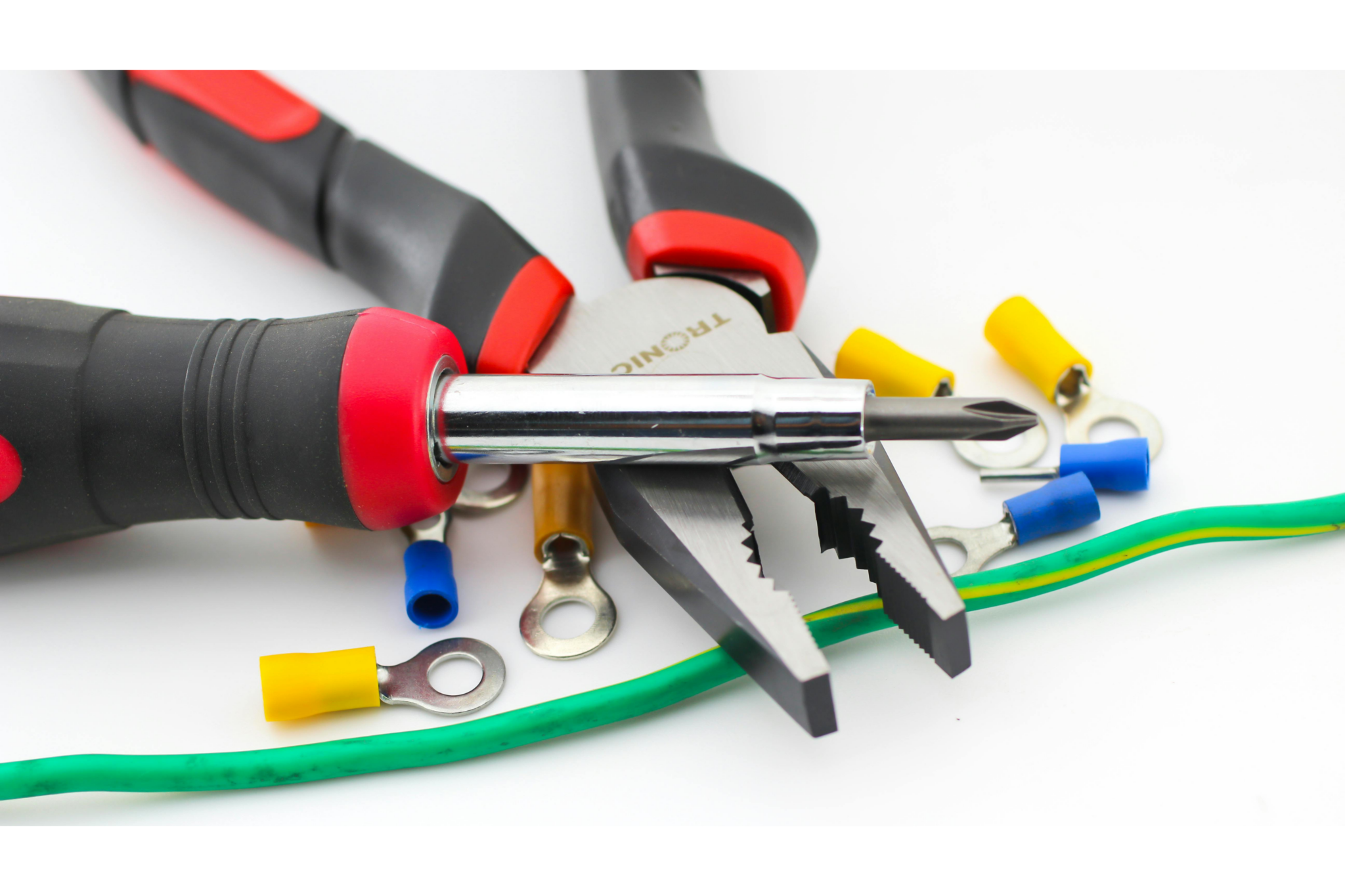 Untitled design (21) "Close-up of electrical tools and components on a white surface, including two pairs of pliers and a Phillips head screwdriver with red and black handles, surrounded by assorted wire terminal connectors in yellow, blue, and silver ring styles, alongside a green and yellow striped electrical ground wire."