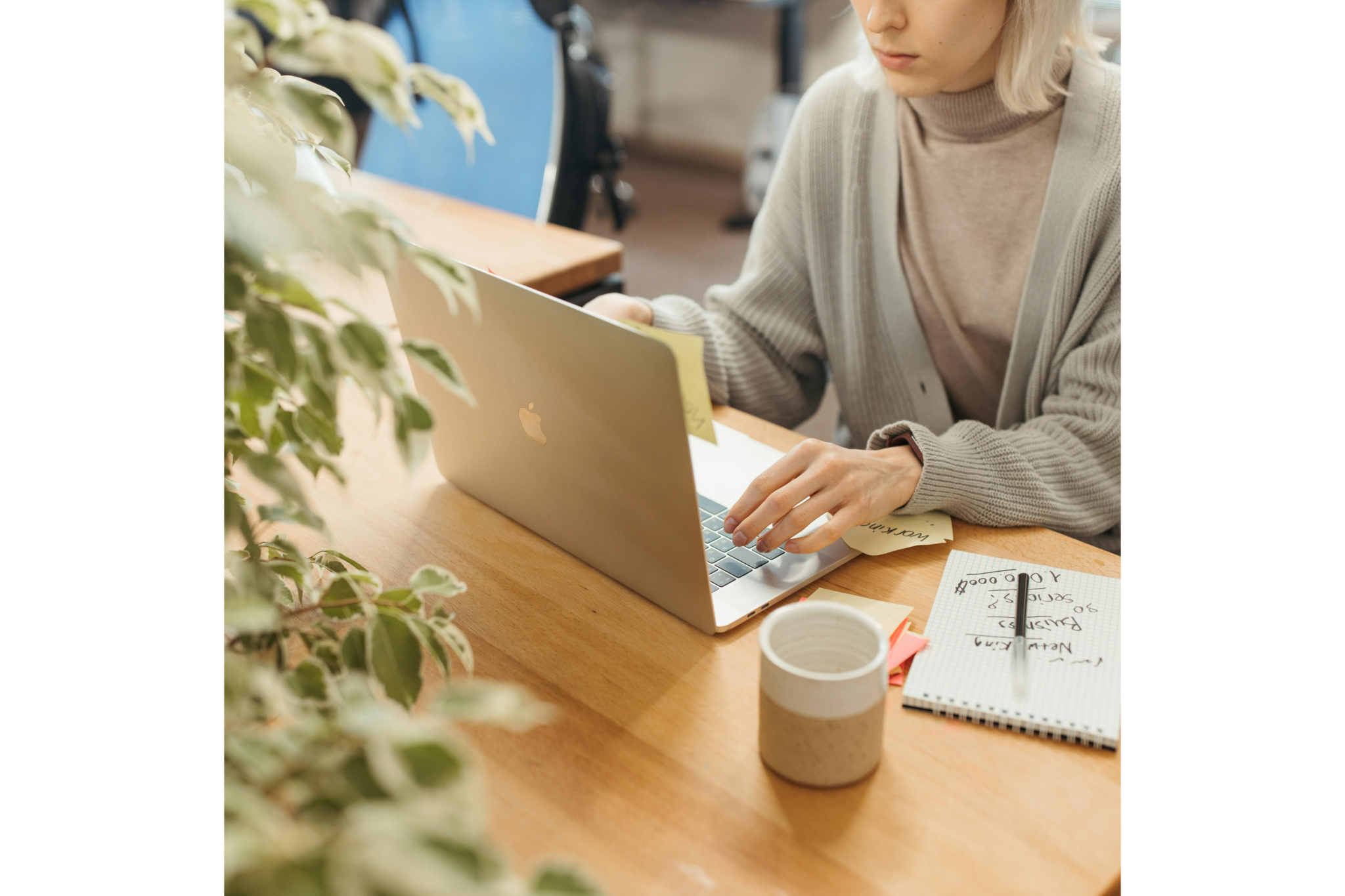 A person with blonde hair wearing a grey cardigan and beige turtleneck working on a silver MacBook laptop at a wooden desk. Surrounding the laptop are yellow sticky notes, a ceramic mug, and a spiral notepad with handwritten business notes and a pen. A leafy green plant is blurred in the foreground.