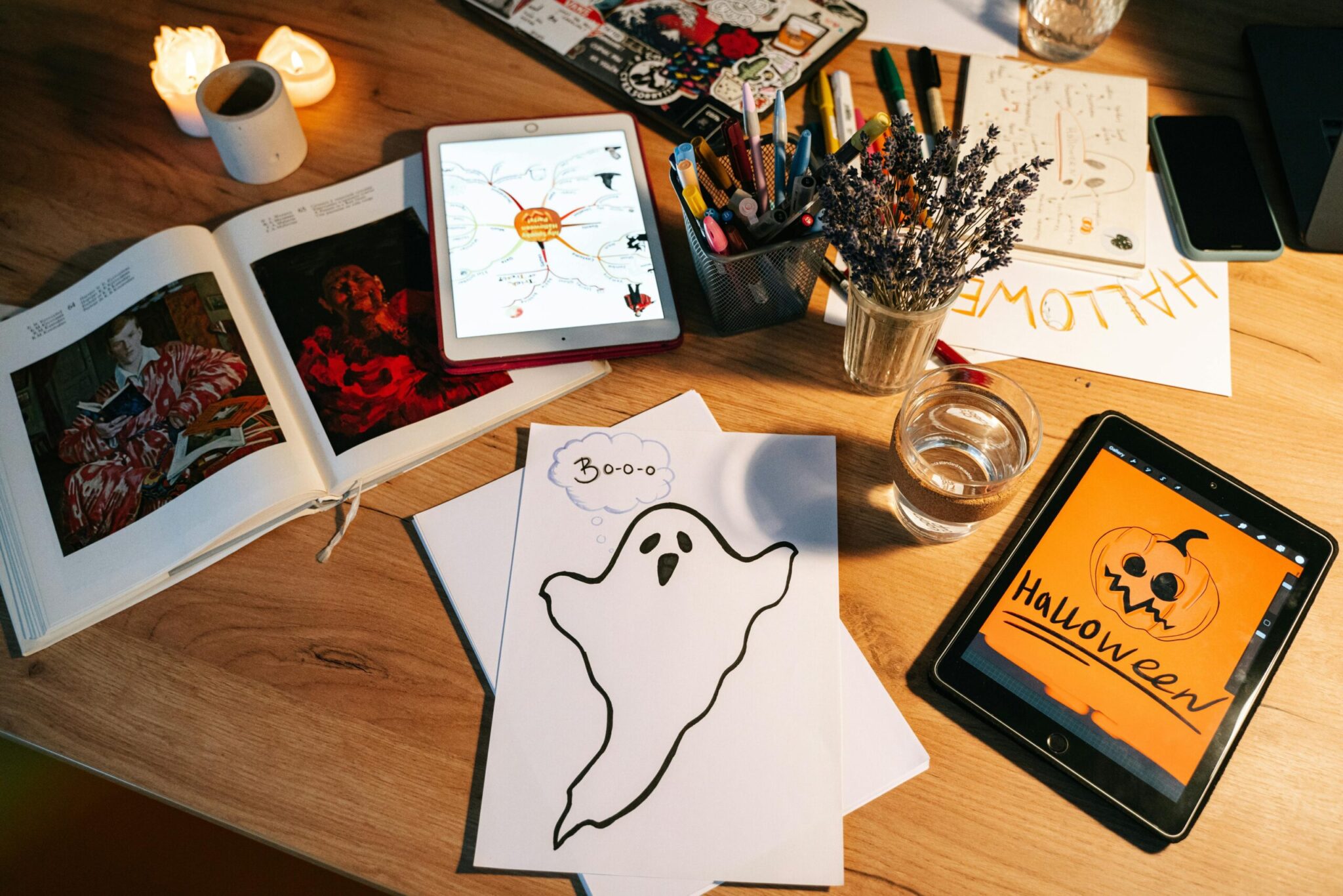 pexels-ivan-s-5428707 Overhead view of a creative workspace with Halloween-themed projects spread across a wooden desk, including a hand-drawn ghost saying 'Boo' on white paper, a tablet displaying a Halloween pumpkin illustration, a second tablet showing a colorful mind map, an open photo book, dried lavender, a glass of water, lit candles, and a cup of markers and pens.